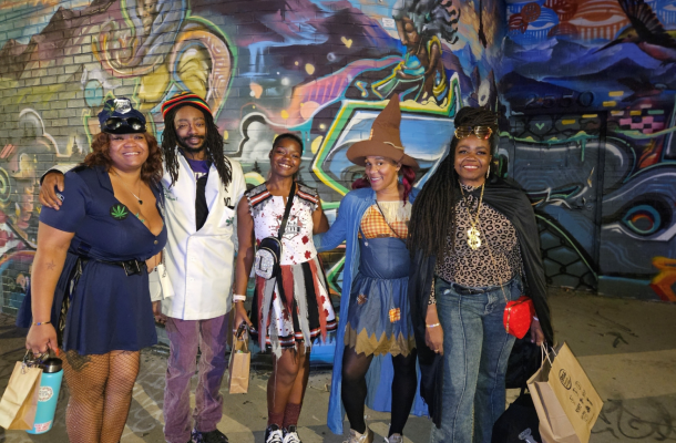 Four Black individuals pose together in their Halloween costumes, radiating creativity and fun. One is dressed as a vampire, another as a witch, the third as a superhero, and the fourth as a ghost. They’re smiling and standing against a festive backdrop with spooky decorations, showcasing their vibrant energy and holiday spirit.