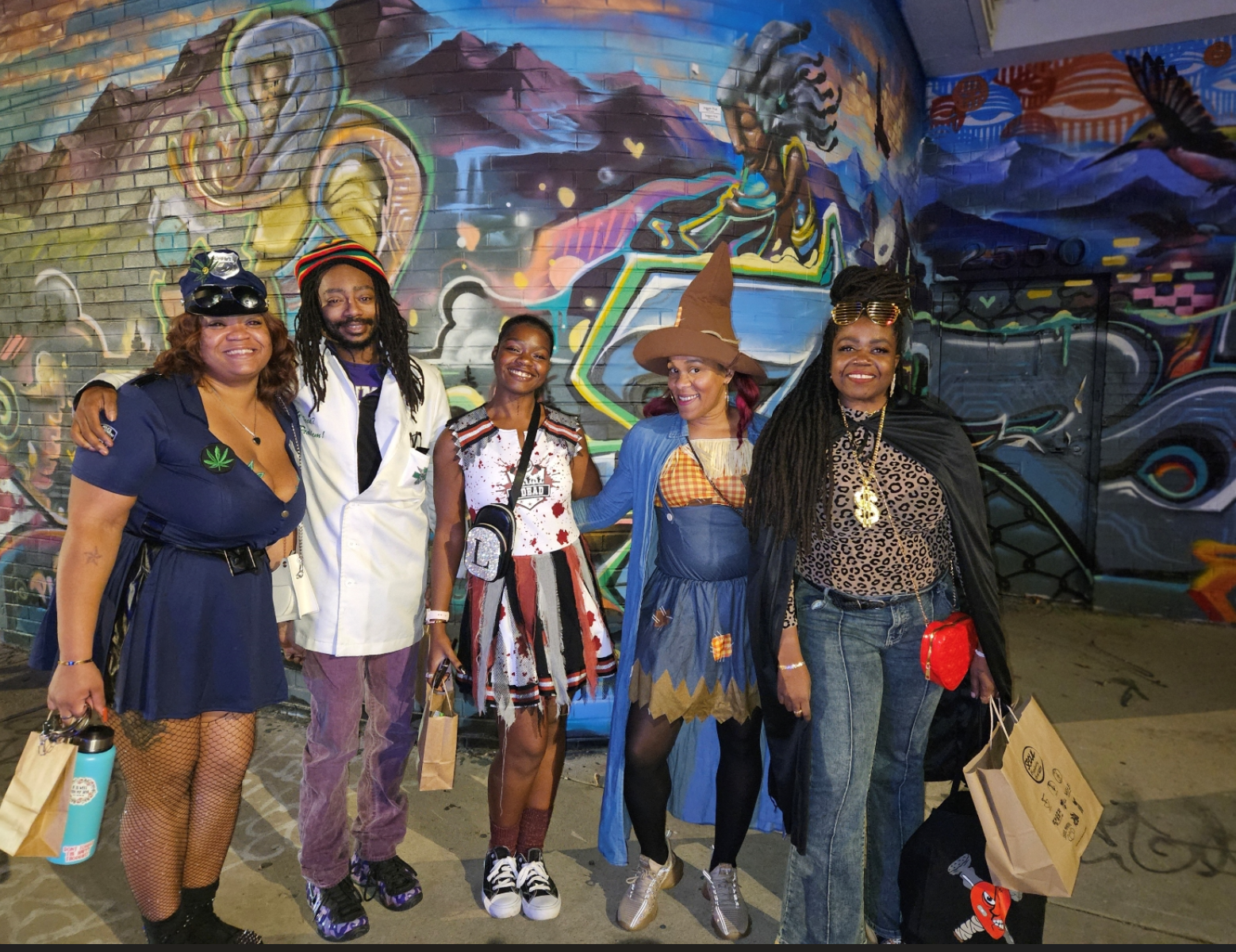 Four Black individuals pose together in their Halloween costumes, radiating creativity and fun. One is dressed as a vampire, another as a witch, the third as a superhero, and the fourth as a ghost. They’re smiling and standing against a festive backdrop with spooky decorations, showcasing their vibrant energy and holiday spirit.