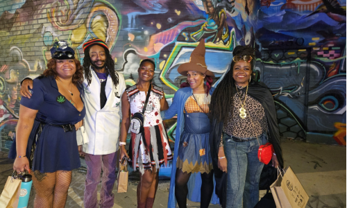 Four Black individuals pose together in their Halloween costumes, radiating creativity and fun. One is dressed as a vampire, another as a witch, the third as a superhero, and the fourth as a ghost. They’re smiling and standing against a festive backdrop with spooky decorations, showcasing their vibrant energy and holiday spirit.